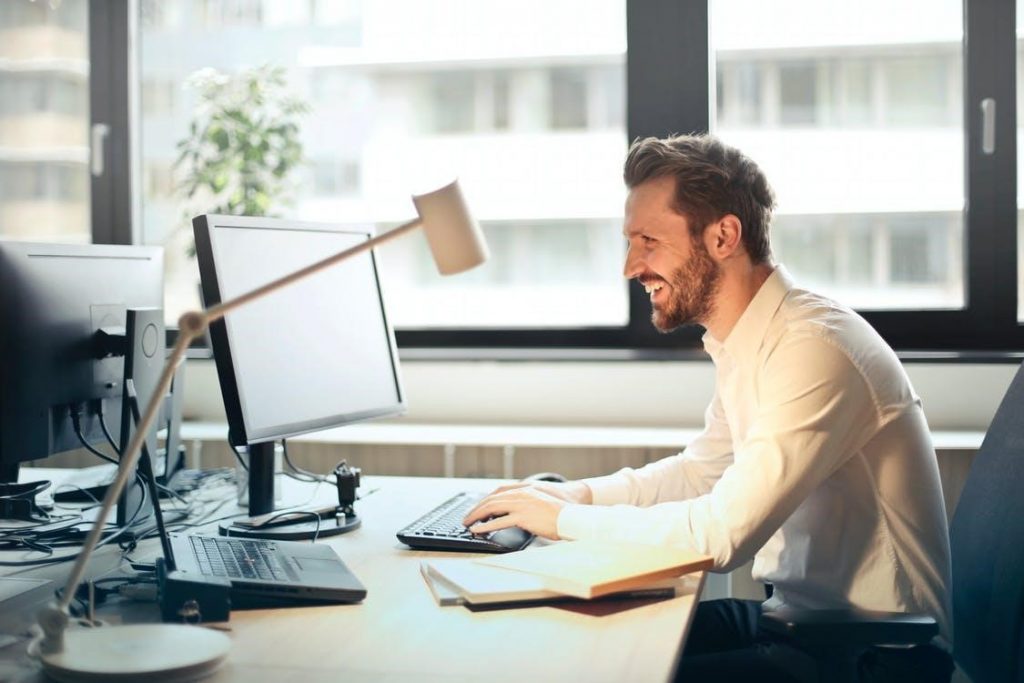 Homem de camisa branca sentado na cadeira preta rolando enquanto enfrenta um computador preto e sorrindo