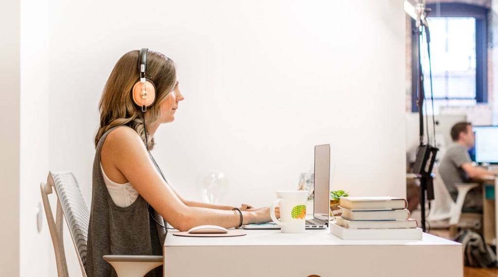 Eine Frau trägt Over-the-Ears-Kopfhörer, während sie an einem Laptop arbeitet. Neben ihr steht eine Kaffeetasse mit dem Bild eines Comic-Igels und ein kleiner Stapel Bücher