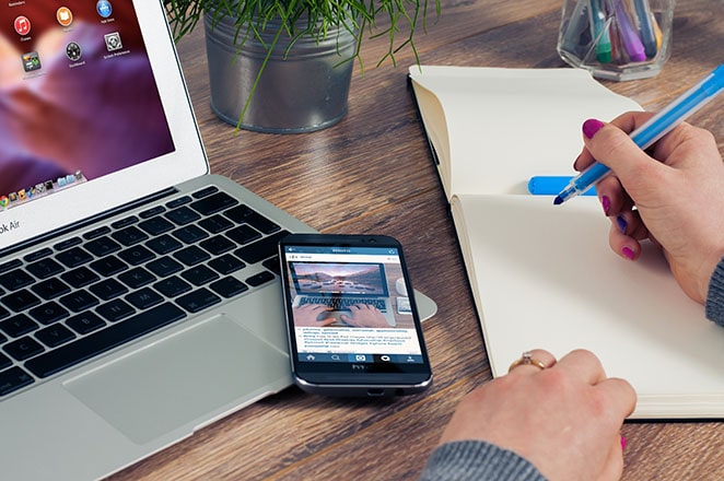 seseorang memegang spidol biru di atas halaman kosong sambil duduk di samping laptop dan smartphone yang terbuka