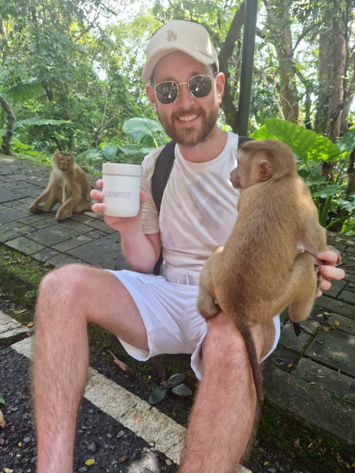 Un uomo sorridente con un berretto da baseball bianco seduto in un ambiente tropicale, con una tazza di bevanda nella mano destra e toccando una scimmia marrone che è in equilibrio sul ginocchio sinistro con la mano sinistra.