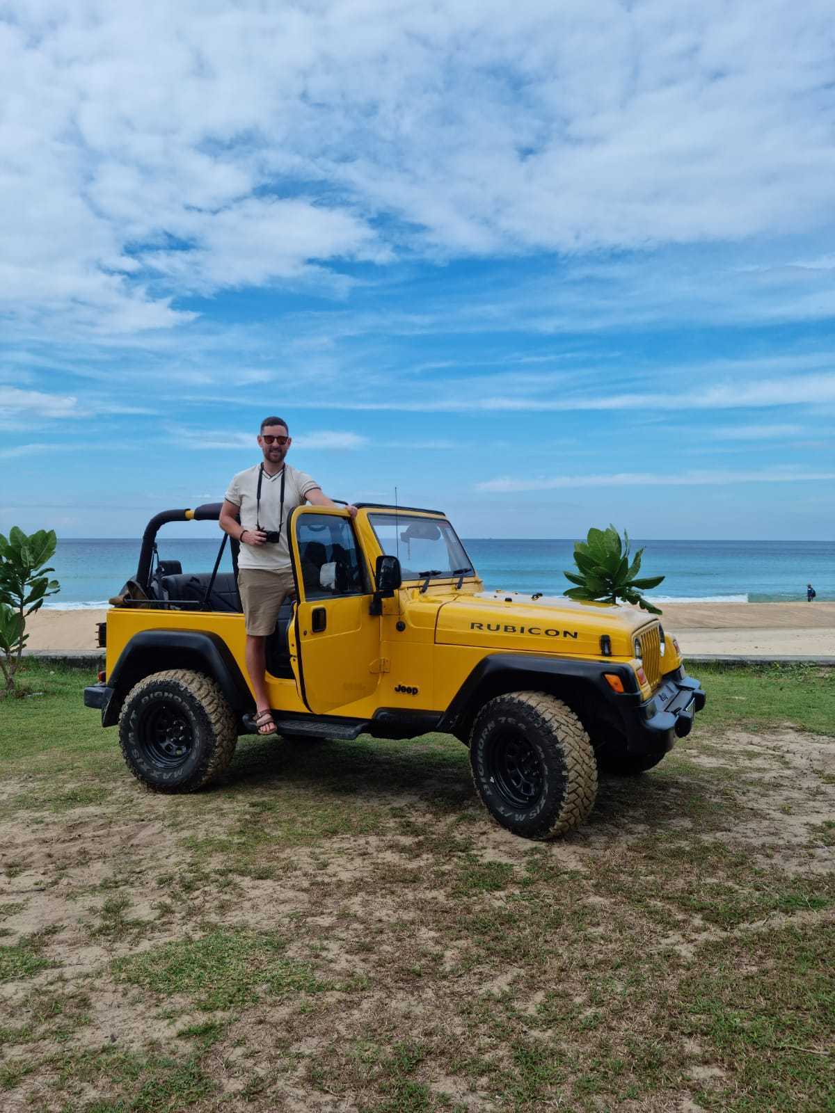 Un uomo in piedi davanti alla porta aperta di una Jeep Rubicon gialla con vista sull'oceano sullo sfondo.