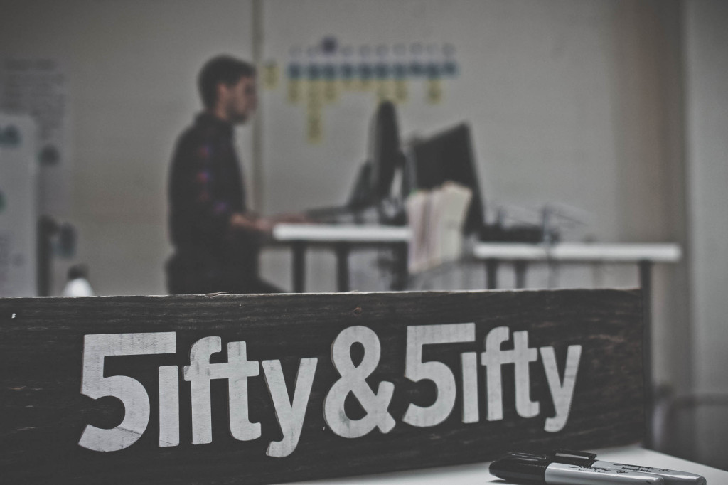 白い文字で「5ifty & 5ifty」と書かれた Fifty & Fifty と書かれた木の板。男はバックグラウンドでコンピューターで作業します