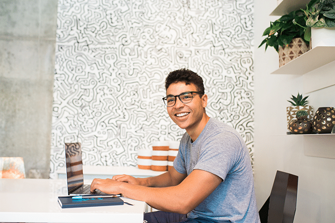 un hombre sonríe a la cámara con las manos listas para escribir en su computadora portátil. Se sienta frente a una pared elaboradamente decorada.