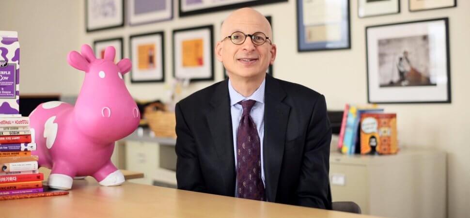 Gambar Seth Godin di mana dia duduk di meja bacanya dengan unicorn dan banyak buku di sekelilingnya